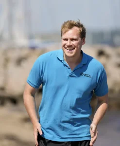 mand smiler ved havn med groen vest i baggrunden