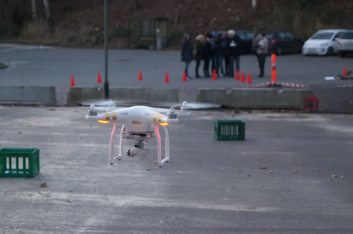Hvid drone flyver over asfalt under teambuilding aktivitet med deltagere samlet i baggrunden