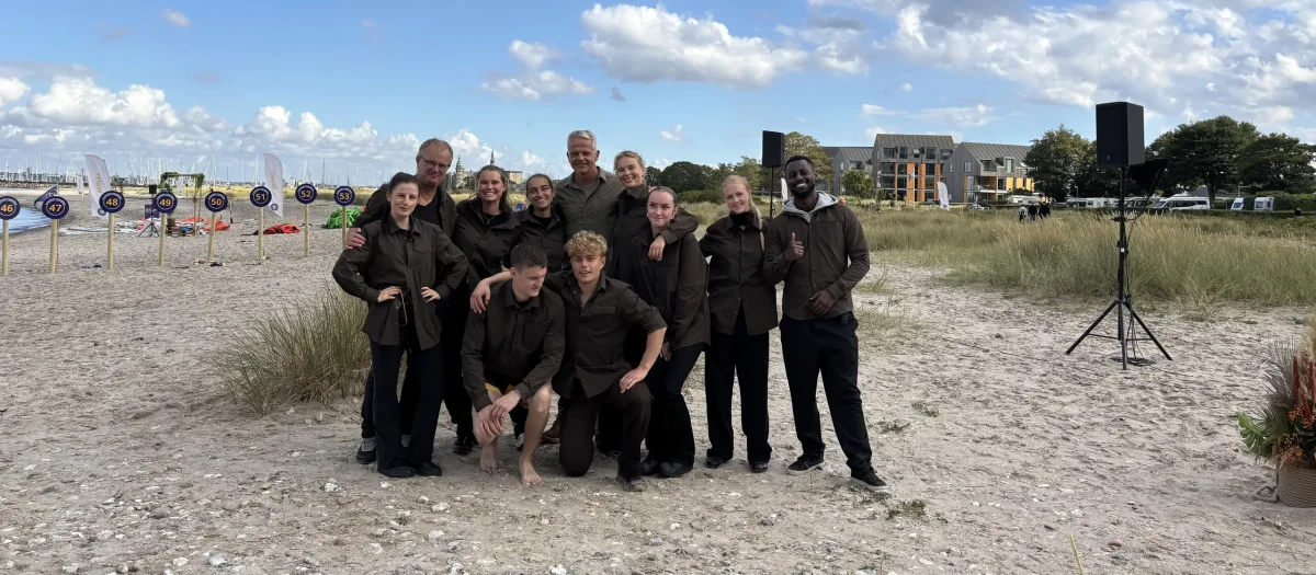 Expedition Robinson Kjelberg deltagere poserer til gruppefoto på sandstrand under blå himmel.