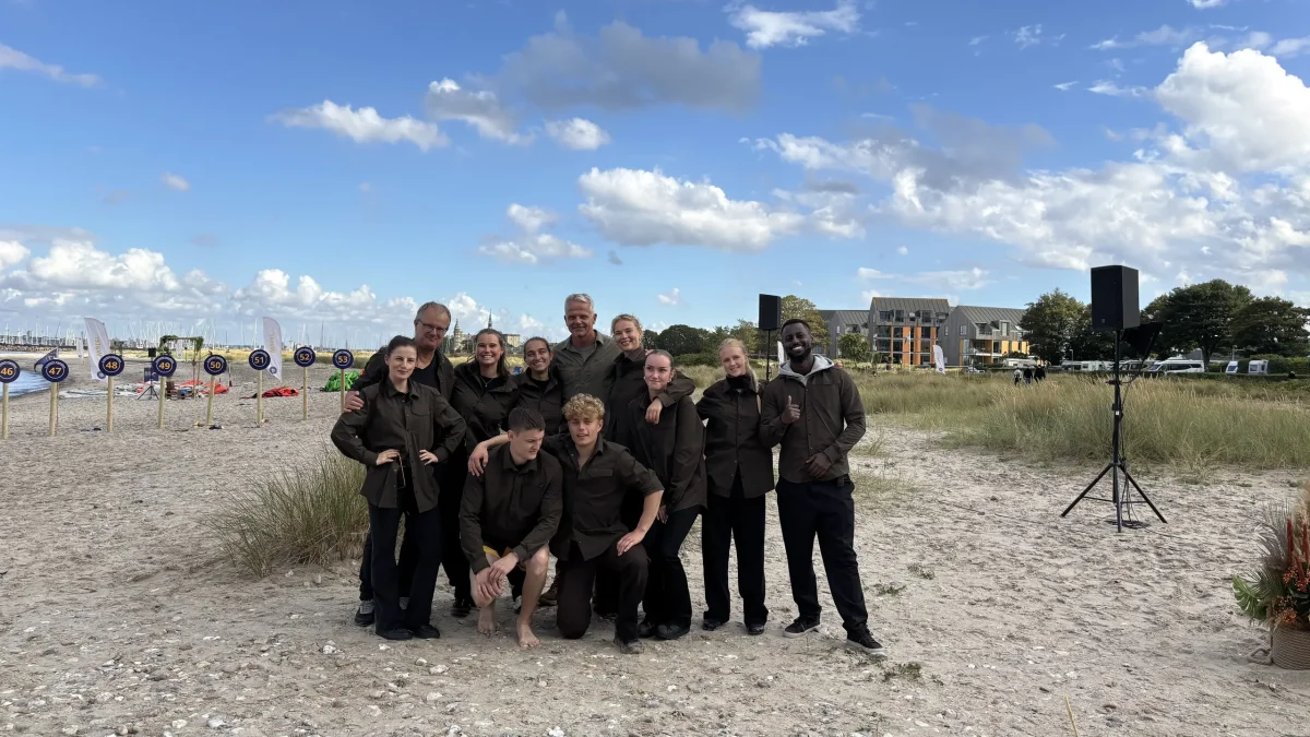 Expedition Robinson Kjelberg deltagere poserer til gruppefoto på sandstrand under blå himmel.