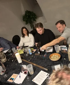 Deltagere i Forræderne spillet samlet rundt om et bord med kort, glas og spilmaterialer under en intens diskussion.
