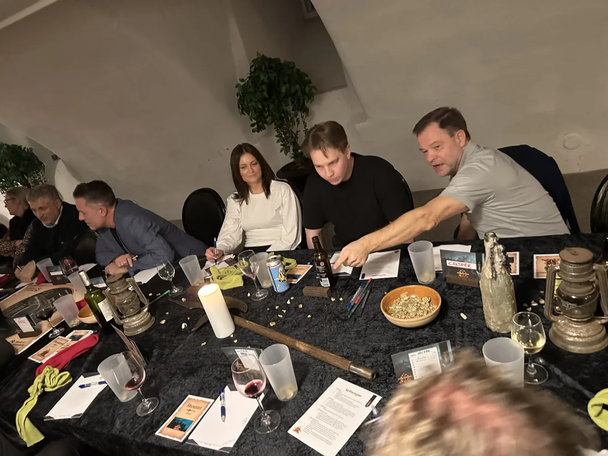 Deltagere i Forræderne spillet samlet rundt om et bord med kort, glas og spilmaterialer under en intens diskussion.