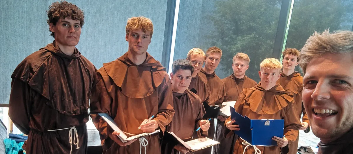 Gruppe af deltagere klædt som munke i brune kutter til Forræderne spillet, der poserer sammen i et lyst lokale.