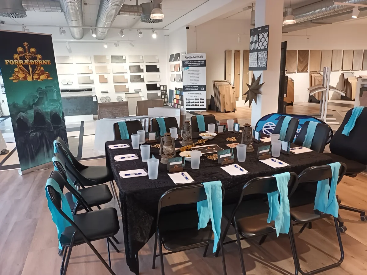 Opstilling til Forræderne spil med sort bord, stole og turkise servietter i showroom miljø.
