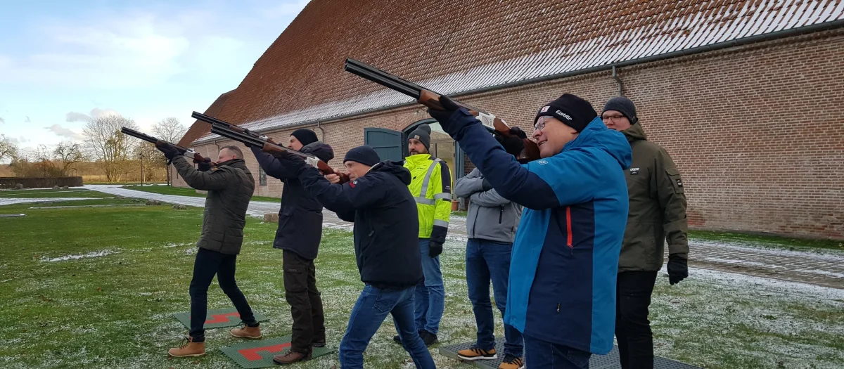 Gruppe personer der øver laser lerdue skydning udendørs foran en stor bygning med instruktør i gul vest.