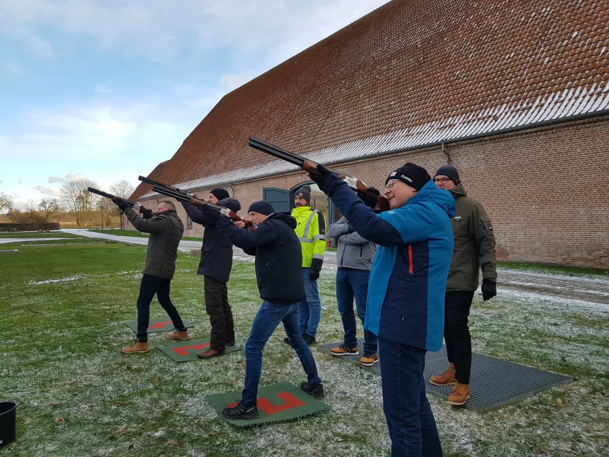 Gruppe personer der øver laser lerdue skydning udendørs foran en stor bygning med instruktør i gul vest.
