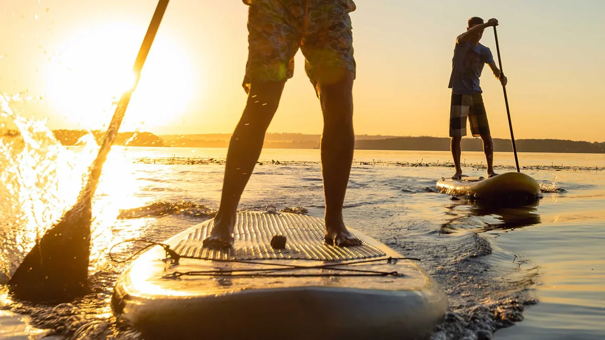To personer står på paddleboards i vandet ved solnedgang med gylden belysning.
