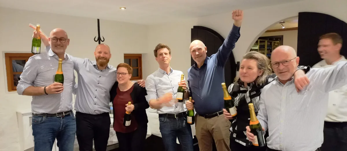 En gruppe mennesker fejrer deres sejr med champagneflasker og jubelråb i et hyggeligt indendørs lokale.