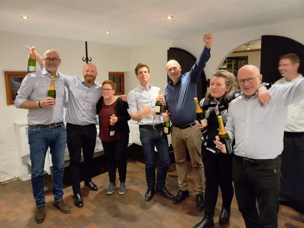 En gruppe mennesker fejrer deres sejr med champagneflasker og jubelråb i et hyggeligt indendørs lokale.
