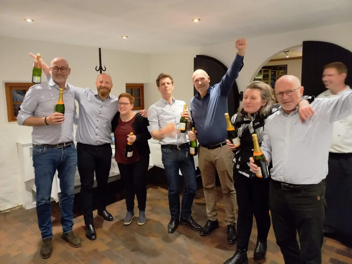 En gruppe mennesker fejrer deres sejr med champagneflasker og jubelråb i et hyggeligt indendørs lokale.