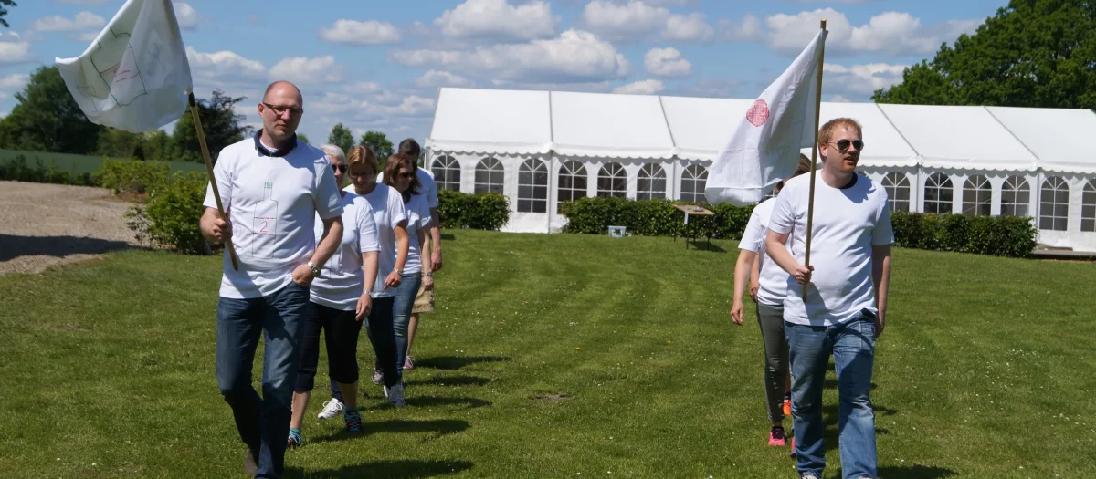 Deltagere i WOA Olympics løber på grøn græsplæne foran hvidt festivaltelt under sommerlig himmel.