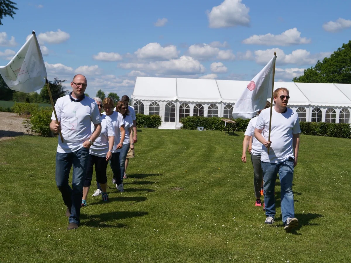 Deltagere i WOA Olympics løber på grøn græsplæne foran hvidt festivaltelt under sommerlig himmel.